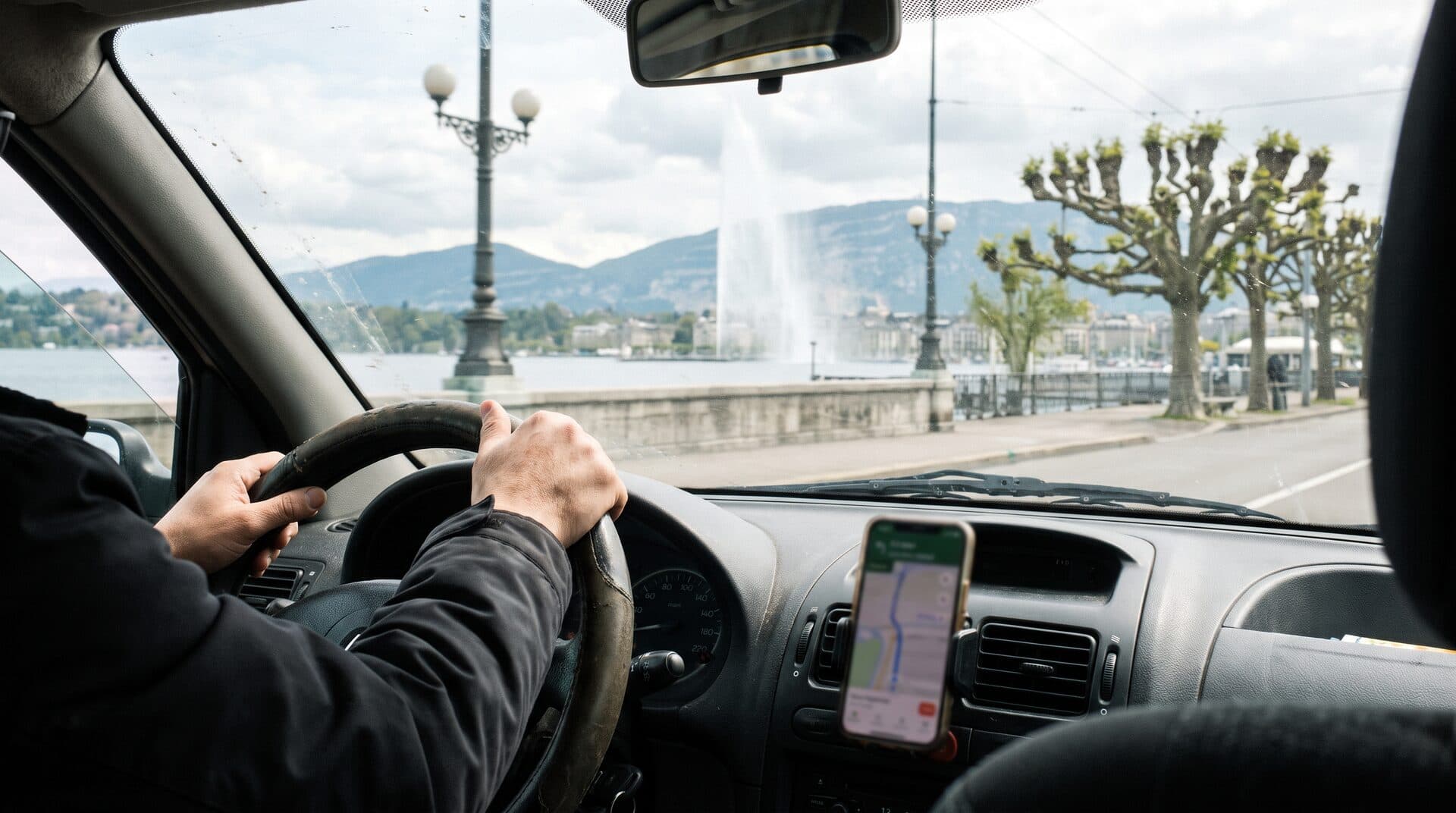Mains d'un chauffeur de taxi sur le volant, quai du Mont-Blanc et jet d'eau de Genève à travers le pare-brise
