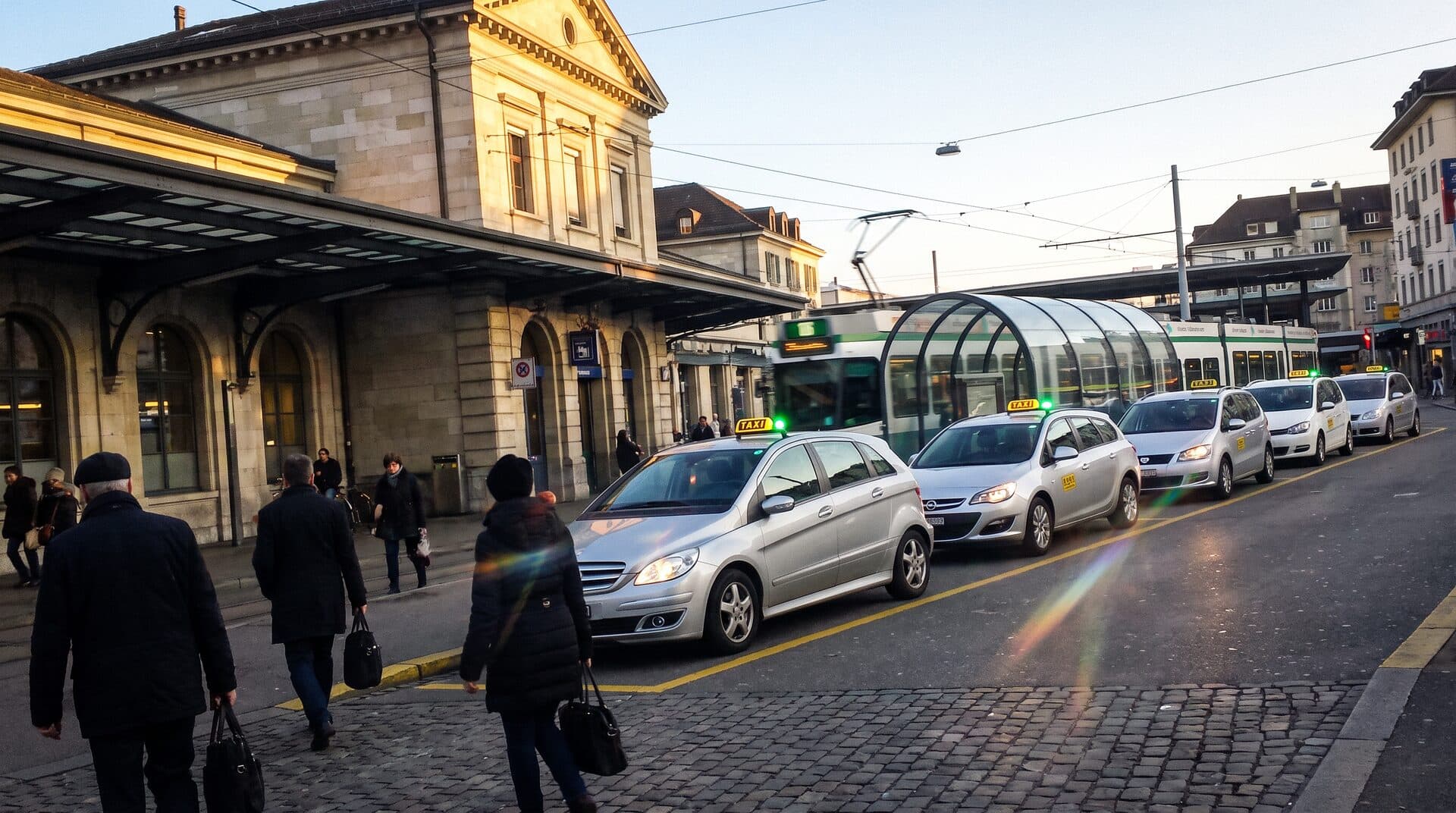 File de taxis devant la gare de Genève-Cornavin en fin de journée, tram et voyageurs au premier plan
