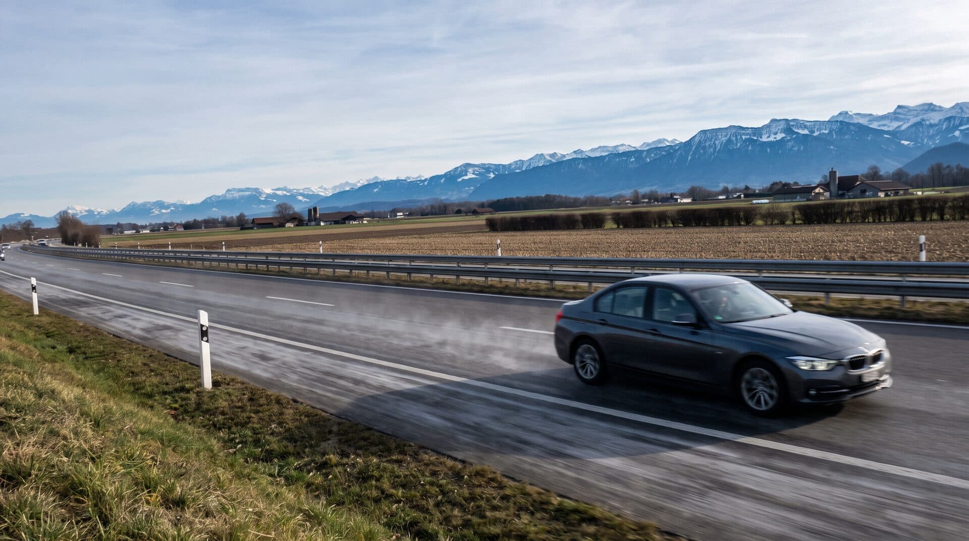 Une berline sombre sur l'autoroute suisse, plaine agricole et chaîne des Alpes enneigées à l'horizon